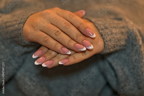 Well-groomed female hands in a grey wool sweater featuring an elegant French manicure with glitter details. Close-up of pastel pink gel nails with almond shape. Soft, cozy autumn and winter fashion.