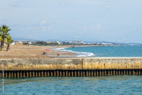 Fototapeta Naklejka Na Ścianę i Meble -  Stone breakwater at a river mouth overlooks a serene beach with flocks of white seagulls, backed by distant resort hotels and hazy mountains under a clear sky. Sorgun, Turkey, Mediterranean.

