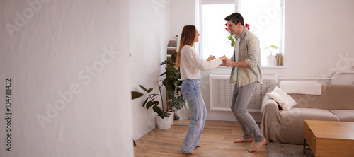 Happy young couple dancing barefoot in a bright cozy living room, holding hands and smiling. Romantic lifestyle moment at home, natural light, modern interior, love and connection concept.