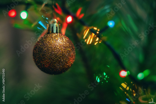 Shiny glitter Christmas ornament hanging on a tree with blurred needles and colorful lights in the background. Symbol of winter holidays, Christmas, New Year, celebration and festive mood.