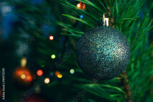 Shiny glitter Christmas ornament hanging on a tree with blurred needles and colorful lights in the background. Symbol of winter holidays, Christmas, New Year, celebration and festive mood.