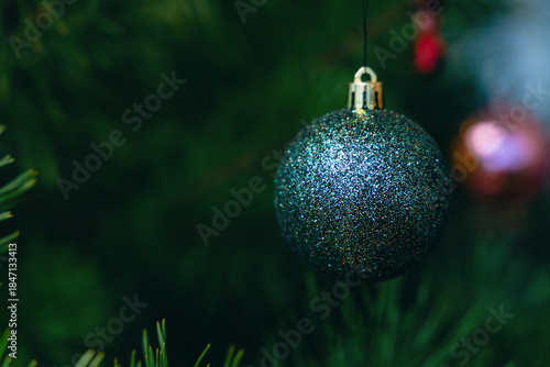 Shiny glitter Christmas ornament hanging on a tree with blurred needles and colorful lights in the background. Symbol of winter holidays, Christmas, New Year, celebration and festive mood.