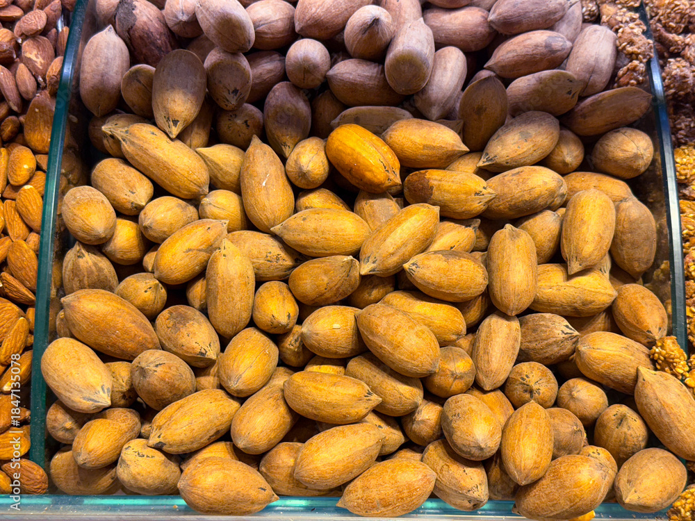 Obraz premium Whole almonds in shells filling a display tray in Istanbul bazaar. Natural nuts, raw ingredients, and nutrient dense food culture expressed through texture, abundance, and agricultural simplicity.