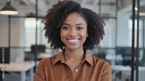 Portrait of happy confident young adult African American business woman standing in office. Smiling professional businesswoman model with curly hair wearing brown shirt looking at camera at work
