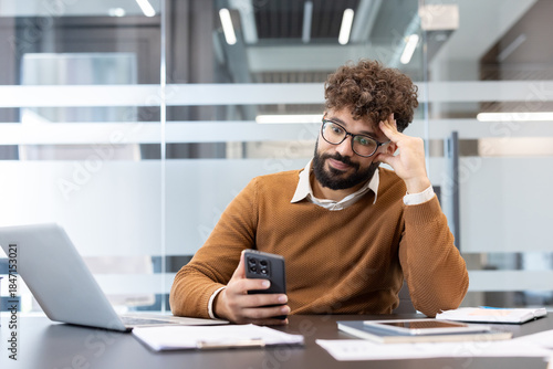 Billede på lærred Young man in glasses sitting at office desk, appearing bored and distracted whil
