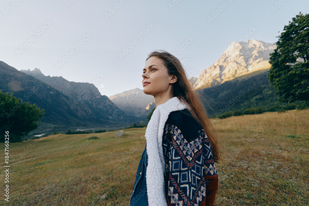 Naklejka premium Woman portrait in mountains meadow, nature outdoors scene with young female in patterned shearling coat, looking in profile at scenic landscape under soft sunlight and peaceful atmosphere