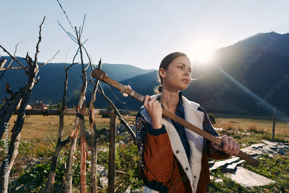 Naklejka premium woman countryside mountains farming field outdoors portrait sun backlit young farmer carrying wooden stick in rustic landscape, thoughtful profile wearing warm jacket and sweater at sunset.