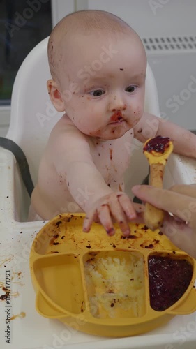 Baby sits in high chair and with appetite eating pureed food with spoon. Emotional messy baby face covered in food. Concept of feeding time and introducing solids to infants. playful exploration food