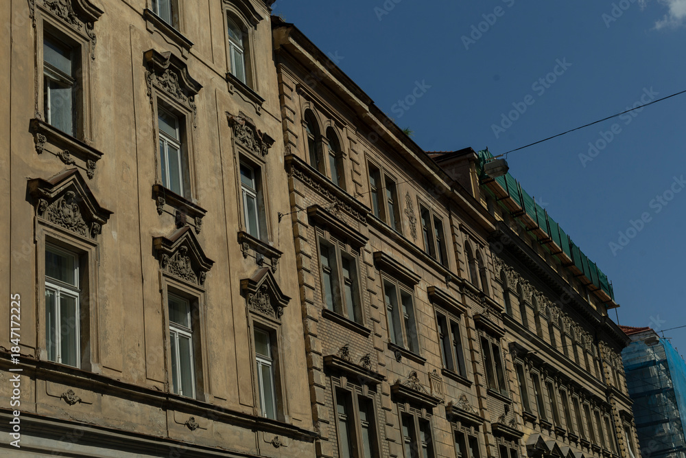 Obraz premium ornate historic facade under clear sky with sculpted pediments, tall sash windows, carved cornices and weathered stucco, warm stone patina, rooftop scaffolding and subtle urban skyline in background