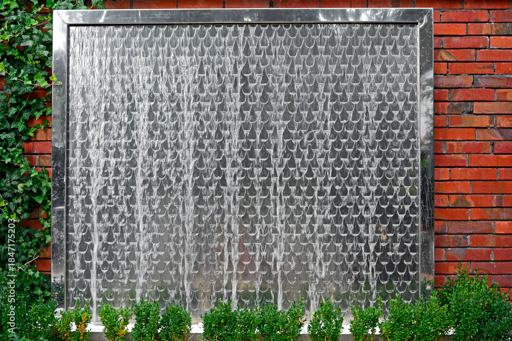 Naklejka premium ściana wodna w ogrodzie. Metalowa ściana wodna na ceglanym murze. water wall in the garden, decorative water fountain cascading in garden, steel wall with a waterfall. Artificial decorative waterfall.