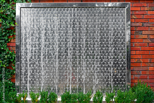 ściana wodna w ogrodzie. Metalowa ściana wodna na ceglanym murze. water wall in the garden, decorative water fountain cascading in garden, steel wall with a waterfall. Artificial decorative waterfall.