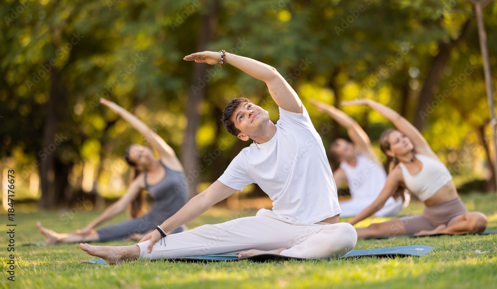 Naklejka premium Sporty young guy in comfortable white sportswear practicing hatha yoga during group class on green lawn in summer park, sitting on mat and doing stretching in Parivrtta Janu Sirsasana asana