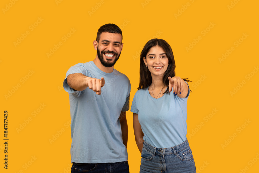 Obraz premium A young Middle Eastern couple stands against a bright yellow background. They are smiling and playfully pointing fingers at the camera, showing excitement and joy in their expression.