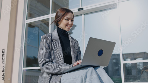 Woman works on laptop outside during daytime at a building with large windows in a casual setting