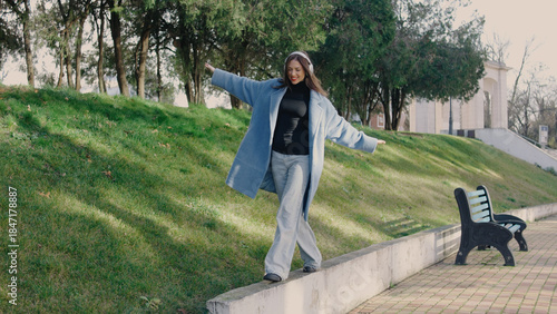 Fotografi Young woman balances on edge of walkway in urban park during sunny day, wearing