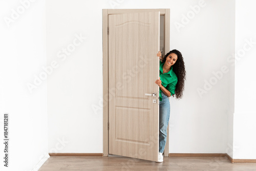 Photography A cheerful young woman stands in the doorway of her modern apartment, smiling and holding the ajar door