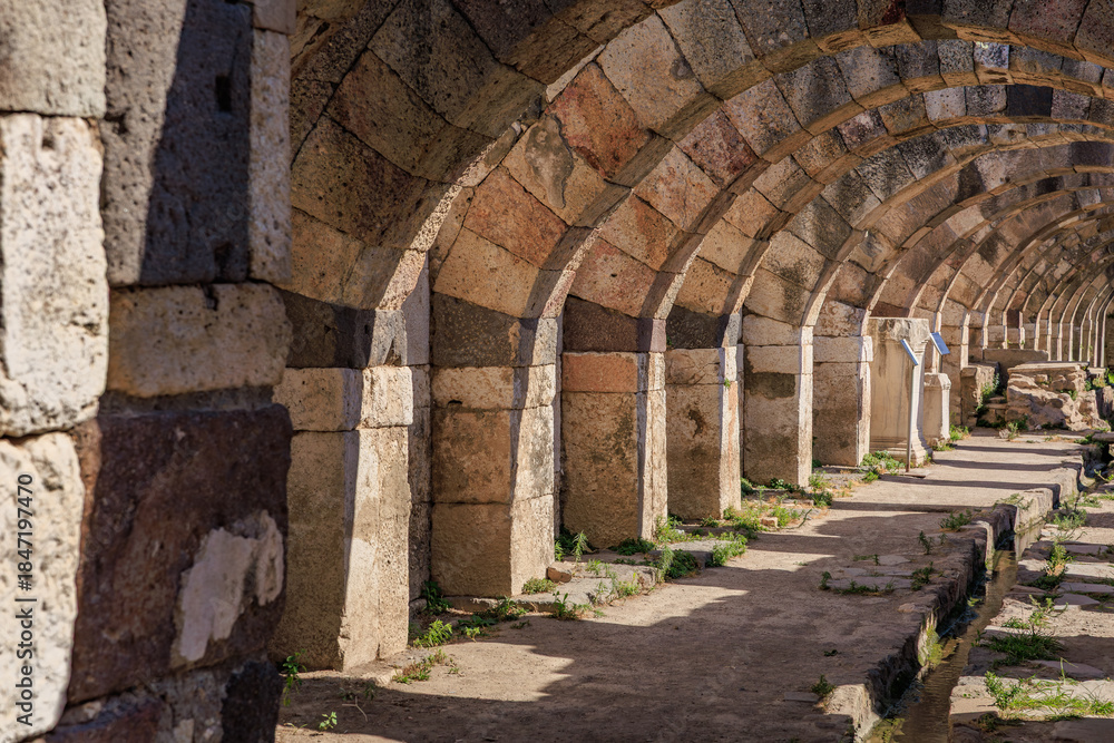 Fototapeta premium Ancient stone archway passageway with sunlit shadows and historical architecture. Authentic ancient Smyrna ruins in Izmir, Turkey