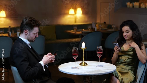 Young couple sitting in the restaurant, having the date while using smartphones. Glasses of wine on the table. Lifestyle, love, relationships concept. Slow motion