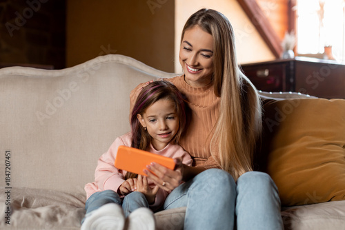 Obraz na plátne A mother and her young daughter sit closely together on a sofa, smiling as they watch a video on a phone