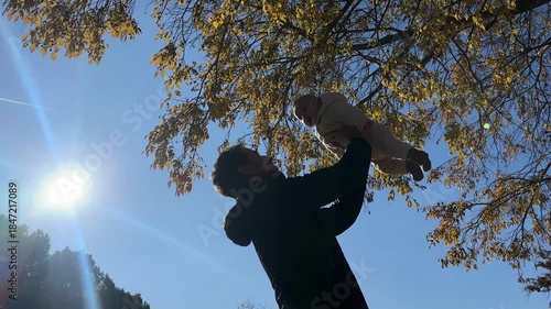 A joyful father lifting his baby into the air as sunlight fills the park