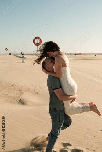 Man lifting woman joyfully on a sandy beach, both smiling in a playful and loving moment under clear skies.