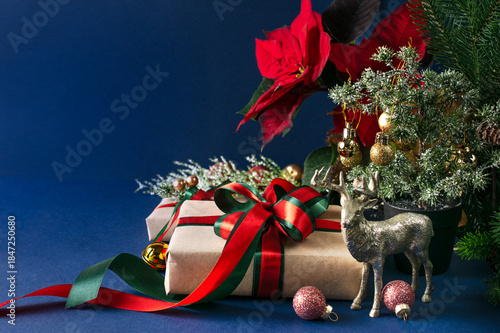 Christmas decor with handmade gift box with bow, red poinsettia, decorative fir branch, deer and balls for tree decoraton on blue wall background. New Year winter holiday composition with presents.