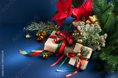 Christmas decor with handmade gift boxes with bow, red poinsettia, decorative fir branch, bells and balls for tree decoraton on blue background. New Year winter holiday composition with presents.