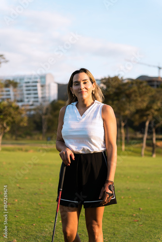 Latin woman golfer in athletic outfit and glove holds club on lush green course, smiling confidently while practicing swing outdoors