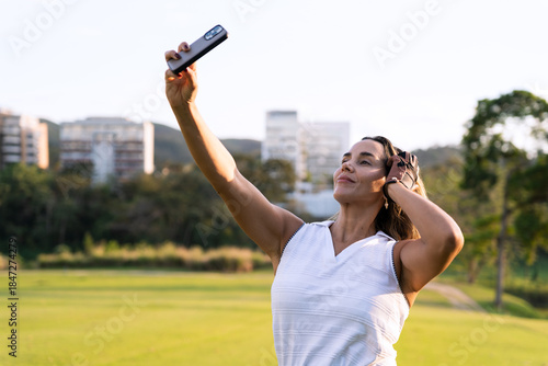 Latin hispanic woman in golf attire taking a selfie with a smartphone on a sunny golf course, enjoying her leisure time outdoors