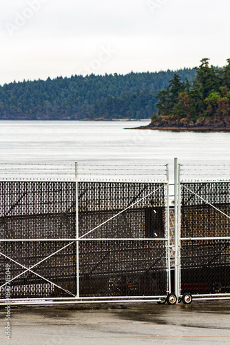Metal Fence Gate By Calm Waterfront With Island Forest In Soft Overcast Light