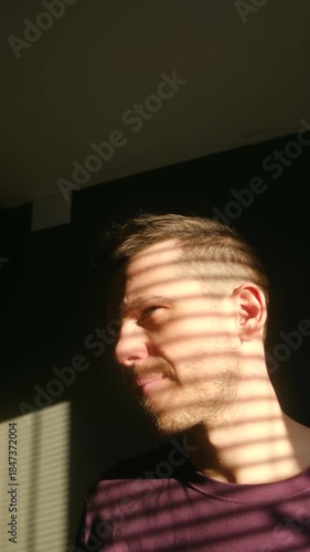 Portrait pensive man touching his hair, his face illuminated by morning sun casting horizontal shadows from blinds, creating dramatic, intimate atmosphere in dark room. Man touching his hair. Vertical