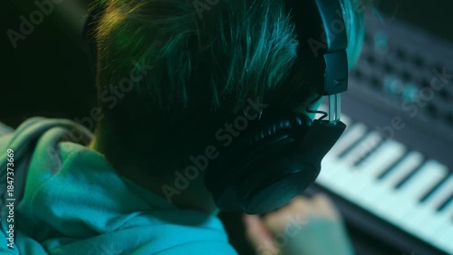 Young talented artist wearing headphones and playing a synthesizer in a dark, neon lit recording studio. Creative process of a musician, dj or composer producing a new song on a keyboard