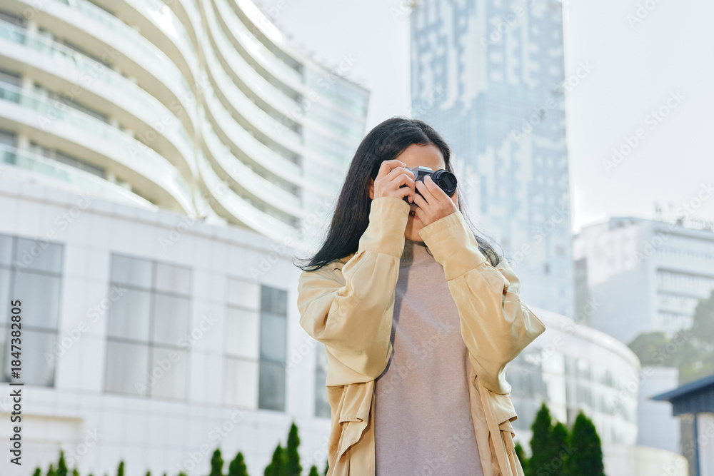 Fototapeta premium Woman with camera adjusting lens for street photography in an urban cityscape, wearing a beige jacket, capturing candid moments among modern buildings and glass towers