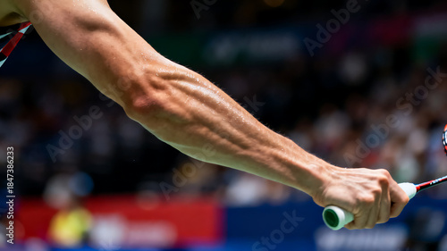 Detailed close-up shot of a badminton athlete’s forearm, showcasing defined muscles and sweat, as they grip a racket mid-action.