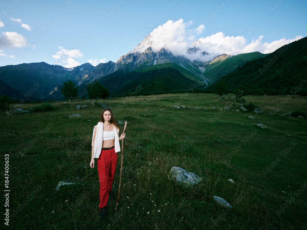 Naklejka premium Woman stands in a green field with a staff, wearing red pants and a white top, mountains in the distance and blue sky behind, depicting outdoor nature and tranquil scenery.