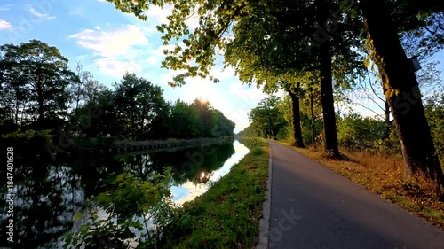 Rijkevorsel, Antwerpse Kempen, Belgium, sunlight through leafy trees by canal vibrant green canopy, shimmering water reflection, paved bike path, warm flare and lens