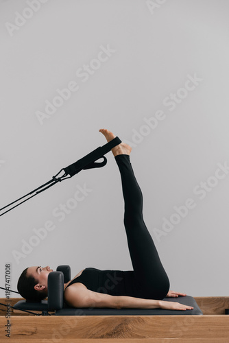 Pilates reformer training machine used in a modern studio as fit caucasian european woman in black leotard performs controlled pilates exercises against a white wall showing fitness lifestyle balance