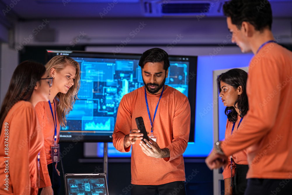 Fototapeta premium A diverse tech event team in matching orange uniforms collaborates in a high-tech control room, coordinating logistics by analyzing real-time data on a tablet and smartphone.
