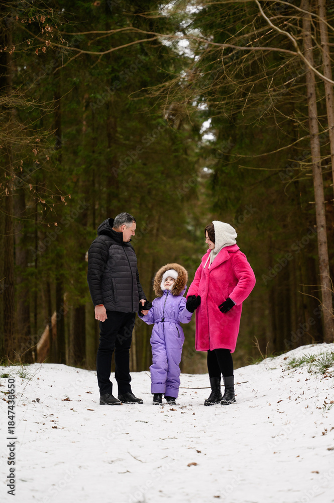 Fototapeta premium Parents and daughter are walking along path in snowy forest, holding hands and looking at each other with love. Family enjoys walk through beautiful winter forest