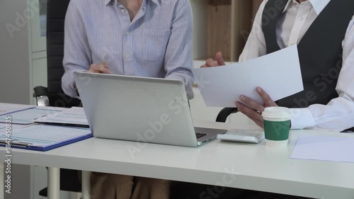 Two Asian business colleagues collaborate in a modern office, reviewing documents and working on a laptop together, showing teamwork, communication, productivity, and professional cooperation