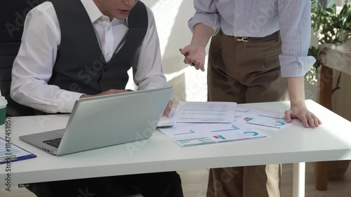Two Asian business colleagues collaborate in a modern office, reviewing documents and working on a laptop together, showing teamwork, communication, productivity, and professional cooperation