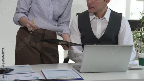Two Asian business colleagues collaborate in a modern office, reviewing documents and working on a laptop together, showing teamwork, communication, productivity, and professional cooperation