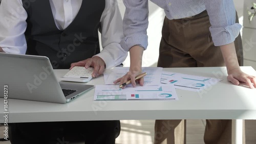 Two Asian business colleagues collaborate in a modern office, reviewing documents and working on a laptop together, showing teamwork, communication, productivity, and professional cooperation