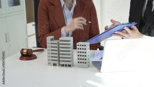 Business professionals discuss real estate and legal matters at an office desk, featuring building models, a judge gavel, and a laptop, symbolizing property law, investment, consultation, corporate