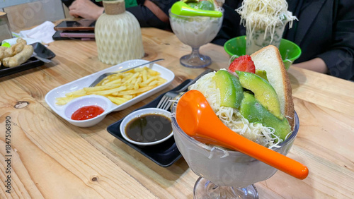 Avocado dessert with grated cheese and strawberry topping, served with toast. French fries with chili sauce and fried snacks with dipping sauce. Sweet drink with cheese topping.