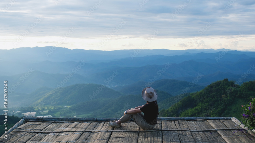 Naklejka premium Rear view of a woman sitting on wooden terrace and looking at a mountain view before sunset