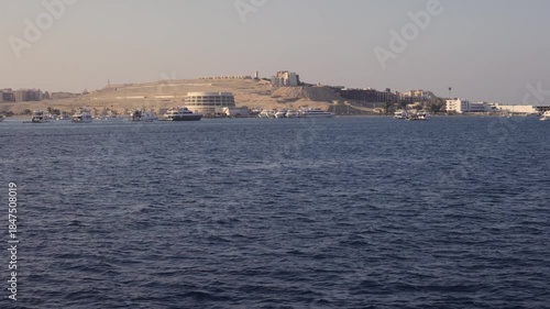 A view of Hurghada's coastal area featuring numerous boats on calm waters and modern buildings along a sandy hill under clear skies.