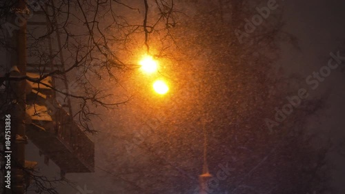Heavy snow falling at night, beautifully illuminated by the warm, orange glow of a city street lamp against a dark winter sky with silhouettes of bare trees, creating a moody atmosphere