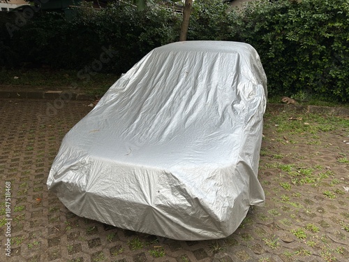 Car covered with silver protective cover parked outdoors on paved surface, protection against weather, dust, rain and sunlight, automotive storage safety concept