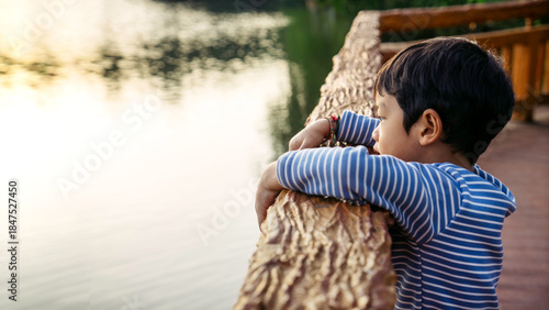 Photos Thoughtful Asian Toddler Leaning on Wooden Railing by Lake, Calm Childhood Momen
